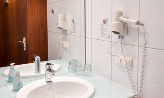 A modern bathroom with a sink, a mirror, and a hairdryer. The walls are tiled, and there is a metal trash can.