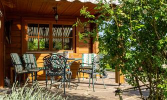 A cozy terrace in front of a wooden cottage with a table and chairs. Surrounded by green plants and plenty of daylight.