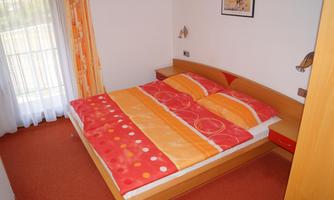 A cozy bedroom with a double bed and colorful bed linen. The walls are light, and a large window provides plenty of daylight.