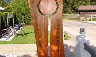 A wooden sign with the inscription "Cyclist Station Sandmore". In the background, trees and a bicycle are depicted.