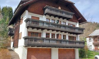 A traditional chalet with wooden cladding and several balconies. It is located in a quiet environment with nature in the background.