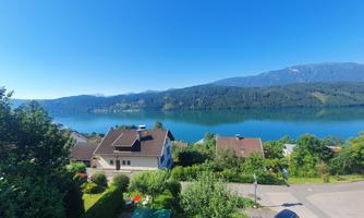 A picturesque landscape with a calm lake and green hills in the background. In the foreground, some houses and trees can be seen.