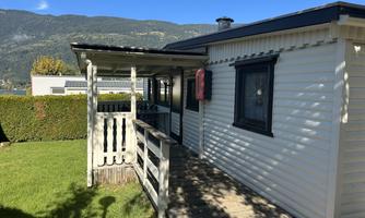 A bright wooden house with a veranda and a well-kept lawn. In the background, mountains and a blue sky are visible.