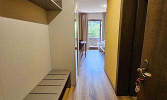 A modern hallway with wooden flooring and bright walls. On the left side, there is a seating area and access to a bright room with windows.