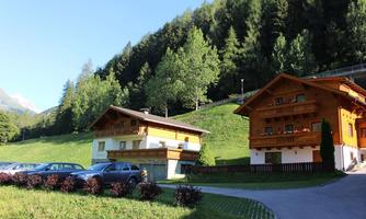 Two charming mountain houses surrounded by green landscape and trees. In the foreground, some cars are parked.