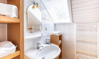 A modern bathroom with a round sink and a large mirror. There is a wooden shelf with towels and space for toiletries.
