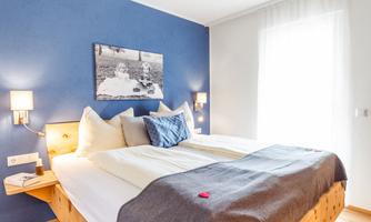 A cozy bedroom with a large bed and blue walls. The room is bright and friendly, with a window that lets in a lot of light.