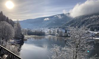 A winter landscape with snow-covered trees and a calm river. The sun shines over the gentle hills.
