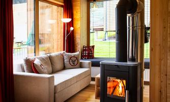 A cozy living space with a comfortable sofa, a wood stove, and large windows. Red curtains and natural wood details create a warm atmosphere.