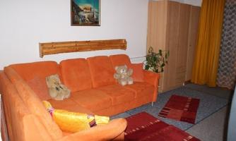 A cozy living room with an orange couch and two plush bears. The room has a light carpet and yellow curtains.