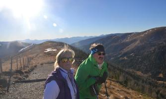 Two people are standing on a mountain path enjoying the view. The landscape is hilly with light snowfields and a clear blue sky.