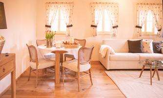 A cozy living room with a dining table, chairs, and a bright sofa. The windows are decorated with curtains, and fresh flowers are on the table.