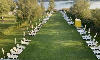 A well-maintained meadow with many sun loungers and sun umbrellas. In the background, a tranquil water flow can be seen.