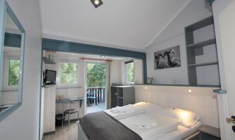 A modern bedroom with a large bed and a desk. There are windows with a view outside and a TV on the wall.