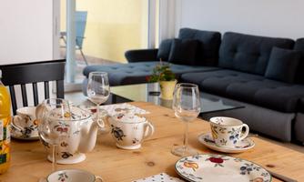 A cozy table set with beautiful dishes and glasses. In the background, there is a modern sofa in a light room.