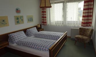 A cozy bedroom with a double bed and an armchair. Large windows let in plenty of light, framed by curtains.