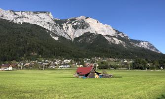 A wide meadow with a small red house in the foreground. In the background, impressive mountains rise and a clear blue sky.
