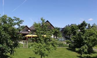 A peaceful garden landscape with green trees and a clear blue sky. In the background, traditional buildings and a terrace with umbrellas can be seen.