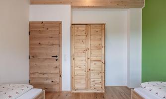 A modern bedroom with two beds and a large wooden wardrobe. The walls are in light tones, with a green accent wall.