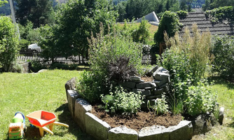 A beautiful garden with a planted bed and plenty of green grass. In the foreground, there is a toy car and a shovel.