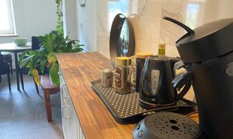 A modern kitchen with a wooden countertop and various kitchen appliances. In the background, a dining table and plants are visible.