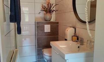 A modern bathroom with white tiles and a large round mirror. A floral decoration and various bathroom items create a pleasant atmosphere.