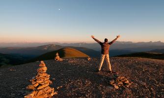 A person is standing on a hill and spreading their arms. In the background, hills and mountains are visible under a clear sky.