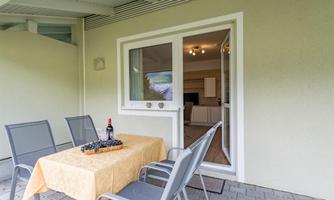 A cozy outdoor seating area with a table and chairs. On the table is a bottle of wine and a fruit basket.
