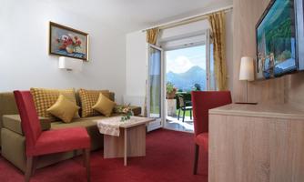 A modern living room with a sofa, red chairs, and a table. Large windows offer a view of the mountains and plenty of daylight.