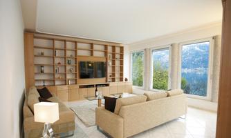 A modern living room with two beige sofas and a glass table. Large windows offer a view of a green landscape and the mountains.