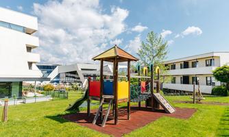 A beautiful playground with a wooden playhouse and slides. The surroundings are green and there are clouds in the blue sky.