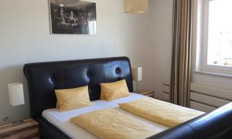 A modern bedroom with a large black bed and bright pillows. The walls are painted light, and there is pleasant lighting from the windows and ceiling lights.