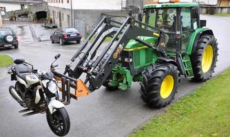A tractor with a loading arm lifts a motorcycle. The scene takes place in a rural setting with houses in the background.
