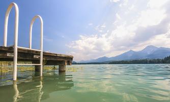 A peaceful lake with a wooden pier in the foreground. In the background, mountains can be seen under a clear blue sky.