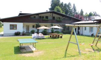 A large house with a beautiful garden. In the foreground are a ping pong table, a swing, and loungers on the lawn.