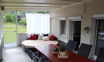 A cozy terrace with a round daybed and a large dining table. In the background, there are green meadows and trees.
