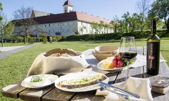 A lovingly set table outdoors with a delicious meal, bread, and wine. In the background, a historic building and a clear blue sky can be seen.