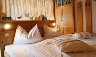 A cozy bedroom with a double bed and decorative pillows. The walls are clad in light wood, and there is a wardrobe in the background.