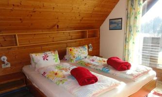 A cozy bedroom with two beds, decorated with colorful pillows and red towels. The wooden ceilings and the large window create a warm atmosphere.