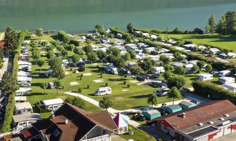 A campsite by the water with many motorhomes and green meadows. In the background, wooded mountains and a tranquil landscape can be seen.