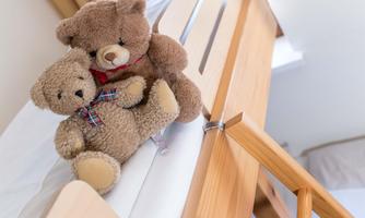 Two plush bears are sitting on the edge of a bed. The larger bear is wearing a red ribbon, while the smaller bear has a checkered scarf.