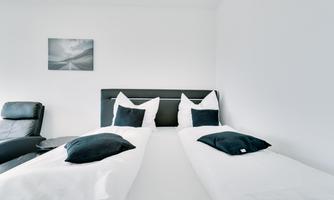 A minimalist bedroom with two single beds and white bedding. A black armchair and a piece of art on the wall complement the decor.