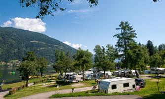 A picturesque campsite by the lake with caravans and trees. In the background, gentle hills and a blue sky are visible.