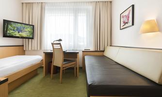 A modern hotel room with a single bed, a sofa, and a desk. The room has large windows with curtains and is bright and inviting.
