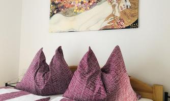 The image shows a bedroom with a bed and large, pointed pillows in purple and white patterns. On the wall hangs a piece of art depicting a woman.