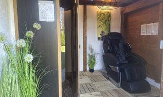 A relaxing wellness area with a massage chair and a sauna cabin. There are also some plants and a cozy wooden paneling visible.