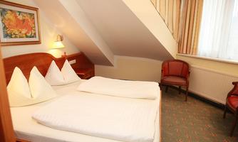 A cozy bedroom with a double bed and two pillows. There is a chair and a window that lets natural light in.