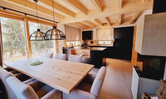 A modern kitchen with wooden ceilings and large windows. The wooden dining table is surrounded by cozy chairs.