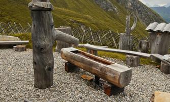 A rustic wooden bench and a tree stump stand on a gravelled area. In the background, green hills and mountains are visible.