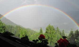 A rainbow stretches over a green mountain landscape. In the foreground, red flowers bloom on a windowsill.
